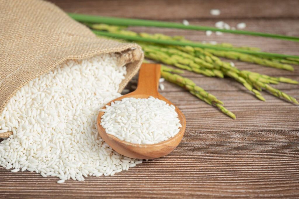 Tips to Reduce High Blood Pressure. A wooden spoon full of white rice sits in front of a burlap sack spilling more rice, with green rice plants on a wooden surface.
