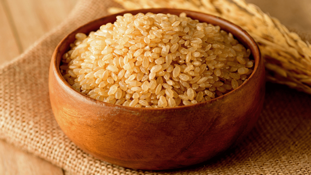 “A wooden bowl full of uncooked brown rice on a burlap cloth, with ears of rice in the background, conveying a sense of harvest and natural, organic produce.