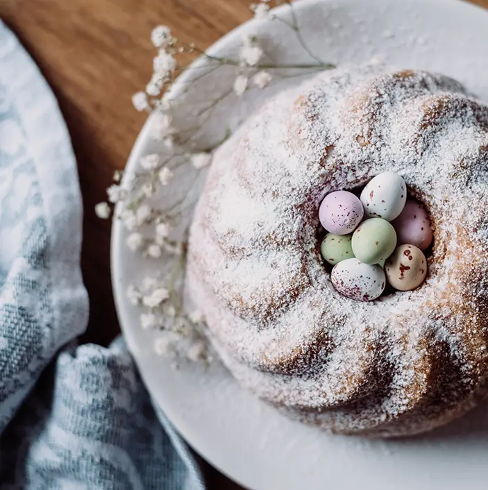 A festive round cake dusted with powdered sugar, adorned with colorful candy-coated chocolate eggs