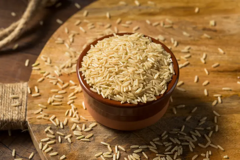 Brown Rice Syrup Market Trends .A brown bowl brimming with uncooked white rice, with some grains scattered around, all bathed in a warm, inviting light.