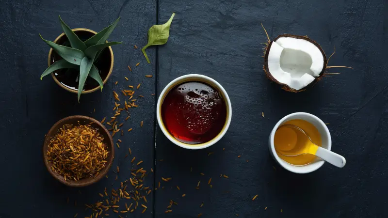 Korean certified rice syrup. A serene flat lay composition featuring wellness items: a cup of tea, a bowl of loose tea leaves, a split coconut, a cup of honey with a spoon, and an aloe vera plant, all arranged on a dark surface