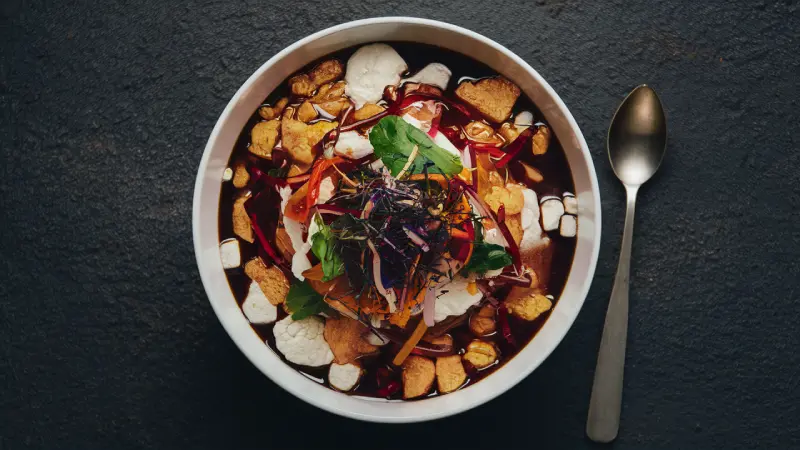 Korean certified rice syrup. A bowl of rich soup garnished with vibrant red chilies, crunchy croutons, and fresh herbs, served on a dark textured surface with a spoon, inviting a comforting and flavorful experience.