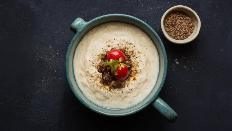 Tapioca Starch Benefits. A top view of a creamy dish in a blue ceramic bowl, garnished with a ripe red cherry tomato with green leaves, and sprinkled with spices. A smaller bowl with ground spices is placed beside it, all on a dark surface.