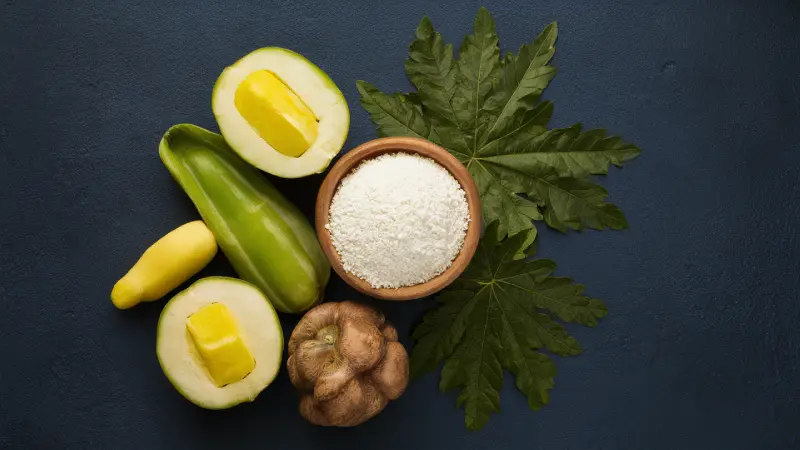Tapioca Starch Benefits. A flat lay of culinary ingredients featuring fresh green bell peppers, yellow and green chili peppers, a bulb of garlic, and a wooden bowl of white grainy substance, possibly salt or sugar, all arranged on a dark blue surface complemented by a large green leaf