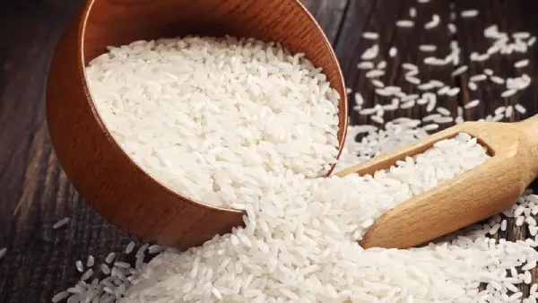 Rice-Based Products. A close-up of a wooden bowl filled with white rice grains, with some grains scattered on a dark textured surface beside a wooden scoop partially filled with rice.