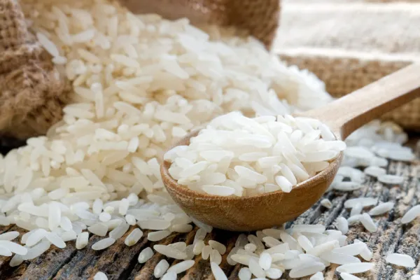 Rice Maltodextrin. Close-up image of a wooden spoon overflowing with white rice grains, resting on a larger pile of rice. The background features a blurred burlap sack and another wooden utensil, highlighting the texture and abundance of the rice.