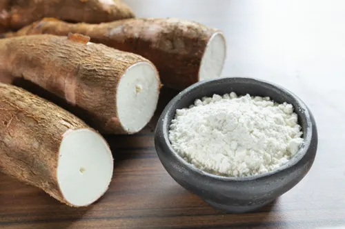 Maltodextrin Vegan. A close-up image of several cylindrical brown cassava roots, with one cut across to show the white interior, alongside a small black bowl filled with white cassava flour, all resting on a wooden surface.