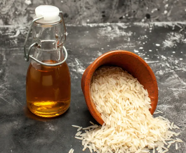 Rice Syrup. A clear glass jar filled with amber-colored liquid, possibly honey or syrup, with a white lid is placed on a dark grey textured surface. Next to it, an overturned brown clay bowl spills uncooked long-grain rice onto the same surface.