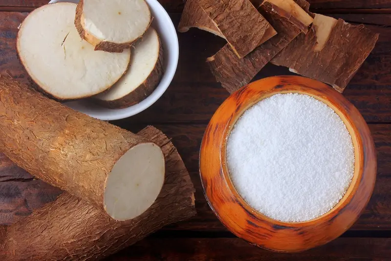 Tapioca Starch. A top-down view of a wooden table with cassava roots, also known as yuca, in various forms. On the left side, there are raw cassava pieces with the bark partially peeled off, revealing the white flesh inside. In the center, there is a small white bowl filled with what appears to be cassava flour. On the right side, there is a round wooden bowl filled with granulated tapioca, which is a product made from cassava. The image showcases the different stages of cassava processing from raw root to edible products.