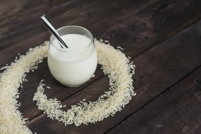 Vegan Milk Weight Loss. An elegant glass of milk with a metal spoon submerged in it, resting on a dark wooden surface. Surrounding the glass is a perfect circle formed by uncooked white rice grains.